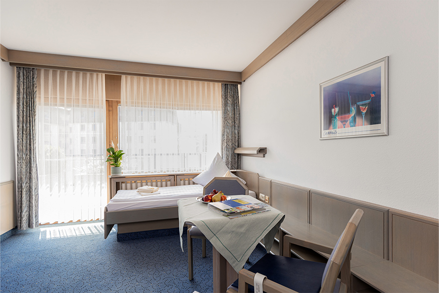 Ein beispielhaftes Patientenzimmer mit großen Fenstern, Balkon, Bett und Sitzecke.